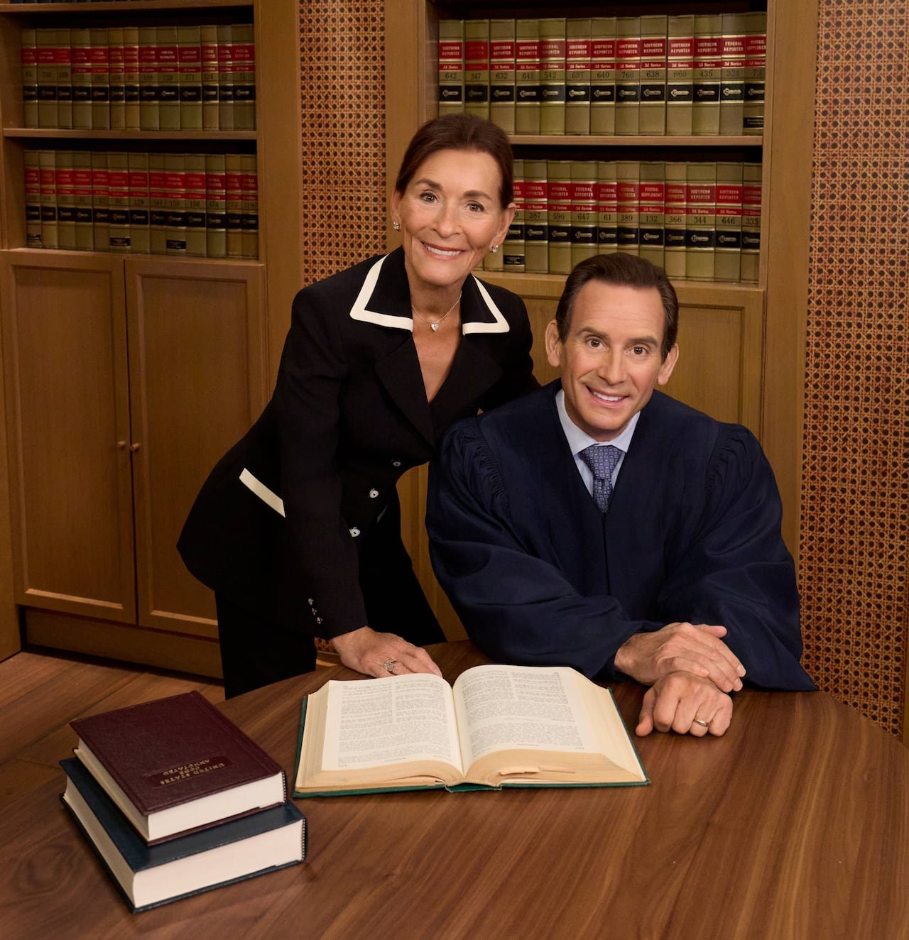 default-131 A woman in a black suit jacket leans over a man in a judge's robe as he's seated at a desk, with a book open in front of him. Both are smiling directly at the camera.
