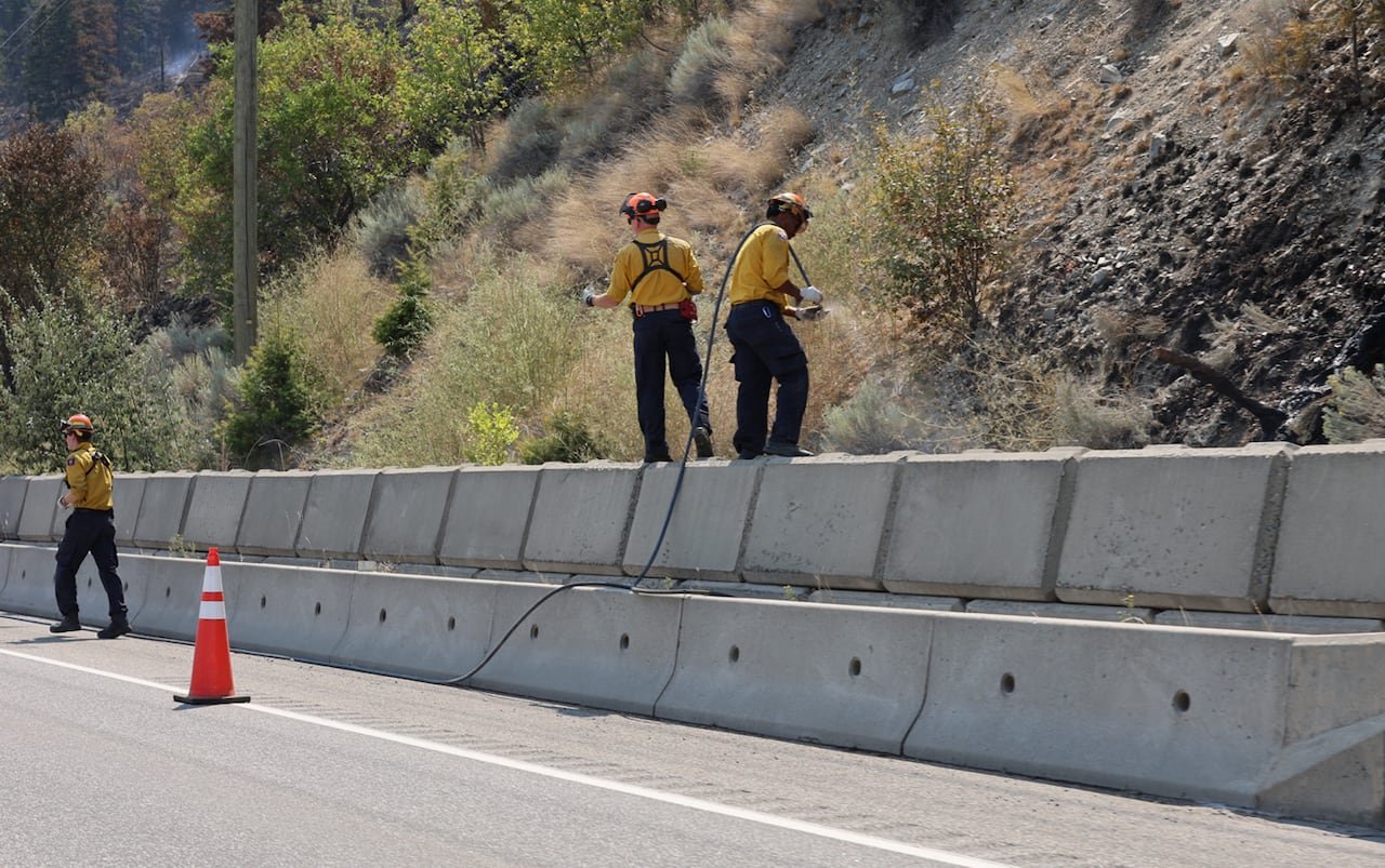 Firefighters wearing yellow are seen on the edge of a highway, with blackened ground. They are using hoses.